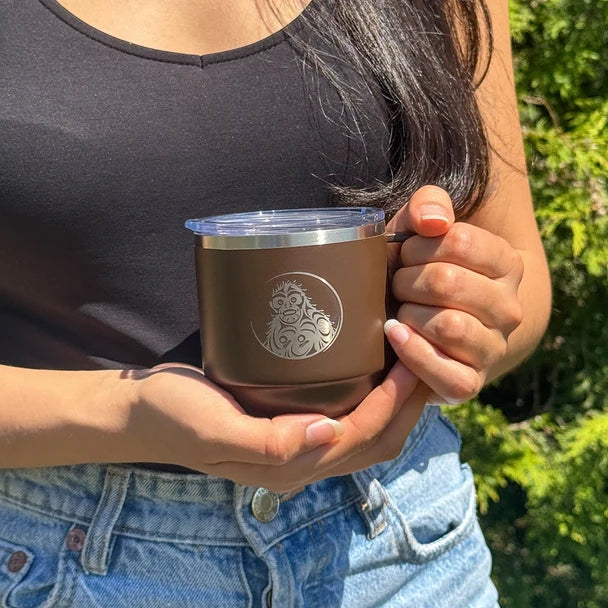Person holding a brown insulated mug with a logo outdoors