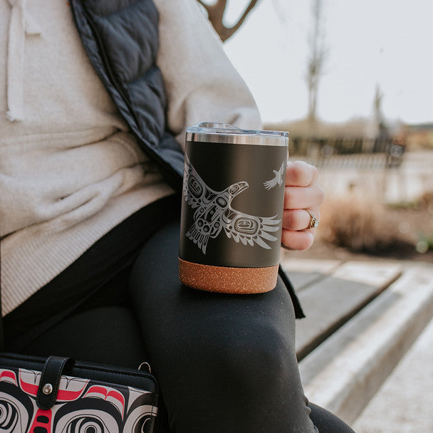 Person holding a black tumbler with a design, sitting outdoors.