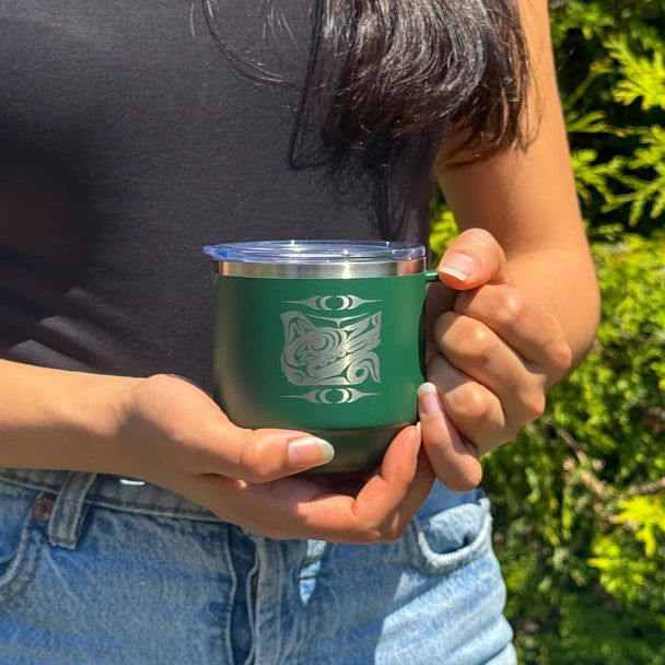 A person outdoors holding a dark green insulated tumbler with a clear lid and a white engraved design. The person is wearing a black top and blue jeans, with green foliage in the background.