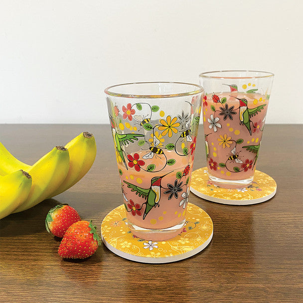 Two floral-patterned glasses on coasters with bananas and strawberries on a wooden surface.